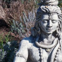 Eine Shiva Statue, sitzt mit geschlossenen Augen in Meditation. Sie ist mit Schmuck und langem Haar geschmückt und steht vor einem Hintergrund aus trockenen Pflanzen und Grün.