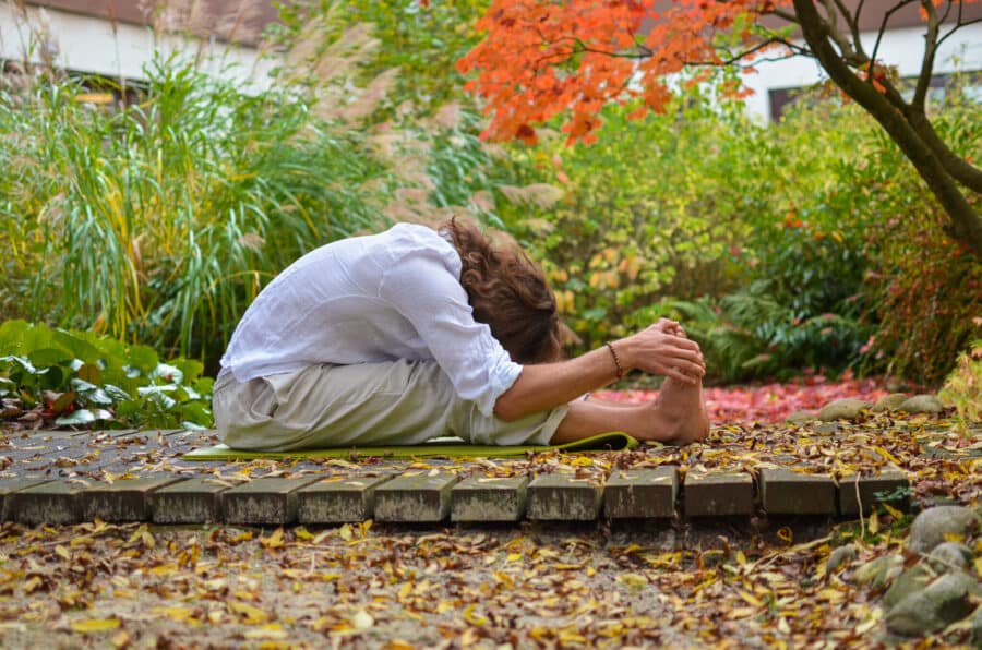 Yoga für die Faszien: Vorwärtsbeuge auf einer Matte im bunten Herbstgarten mit gerundetem Rücken