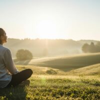 Person sitzt aufrecht im Gras und meditiert bei Sonnenaufgang in weiter Natur
