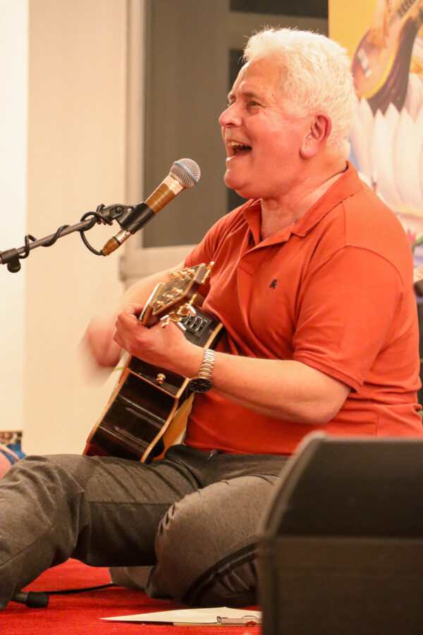 Eine Person mit kurzen grauen Haaren, einem orangefarbenen Hemd und grauen Hosen sitzt auf einem roten Teppich und spielt Akustikgitarre. Sie singt in ein Mikrofon und wirkt fröhlich und in ihre Darbietung vertieft.
