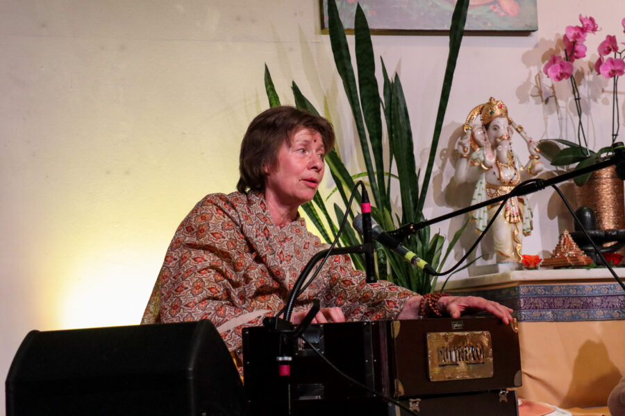 Eine Frau in gemusterter Kleidung spielt Harmonium, umgeben von Pflanzen und einer Götterstatue. Rosa Orchideen zieren den Hintergrund. Sie singt in ein Mikrofon, konzentriert und versunken in die Musik.