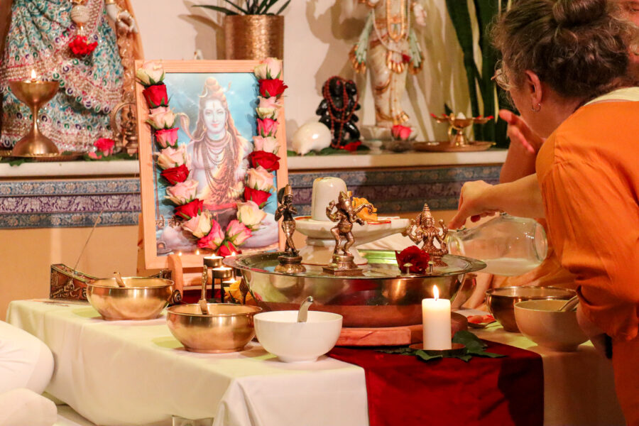 Eine Gruppe von Menschen sitzt um einen Tisch, der für ein hinduistisches Ritual gedeckt ist. Auf dem Tisch stehen ein gerahmtes Bild einer Gottheit, kleine Statuen, Schalen und eine brennende Kerze. Eine Frau gießt Wasser in eine Schale. Der Tisch ist mit Blumen dekoriert.