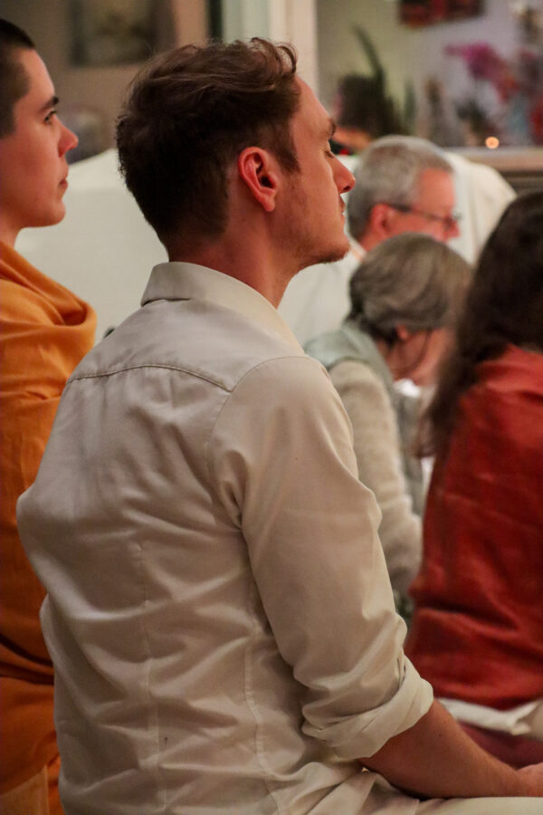 Ein Mann in weißem Hemd sitzt im Profil in einem Innenbereich, wo andere einer meditativen oder kontemplativen Tätigkeit nachgehen. Gedämpftes Licht und weicher Fokus schaffen eine ruhige Atmosphäre. Andere Personen sitzen in der Nähe und tragen zur ruhigen Atmosphäre bei.
