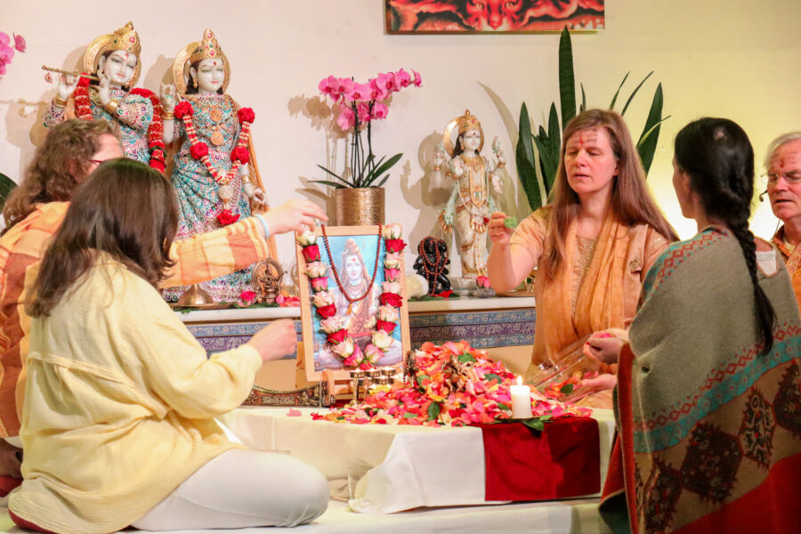 Eine Gruppe von Menschen in traditioneller Kleidung sitzt um einen Tisch mit Blumen und einer brennenden Kerze. Sie stehen vor religiösen Statuen und halten ein gerahmtes Bild, das mit einer Girlande geschmückt ist. Die Szene scheint Teil einer spirituellen oder religiösen Zeremonie zu sein.