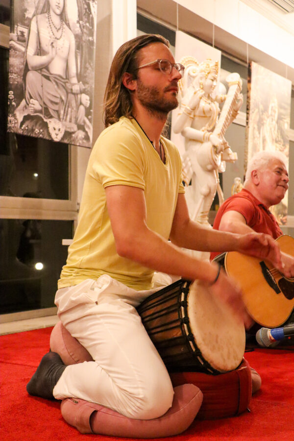 Eine Person mit langen Haaren und Brille, die ein gelbes Hemd und weiße Hosen trägt, spielt auf dem Boden kniend eine Trommel. Eine andere Person in einem roten Hemd spielt im Hintergrund Gitarre. Dahinter sind Statuen und Kunstwerke ausgestellt.