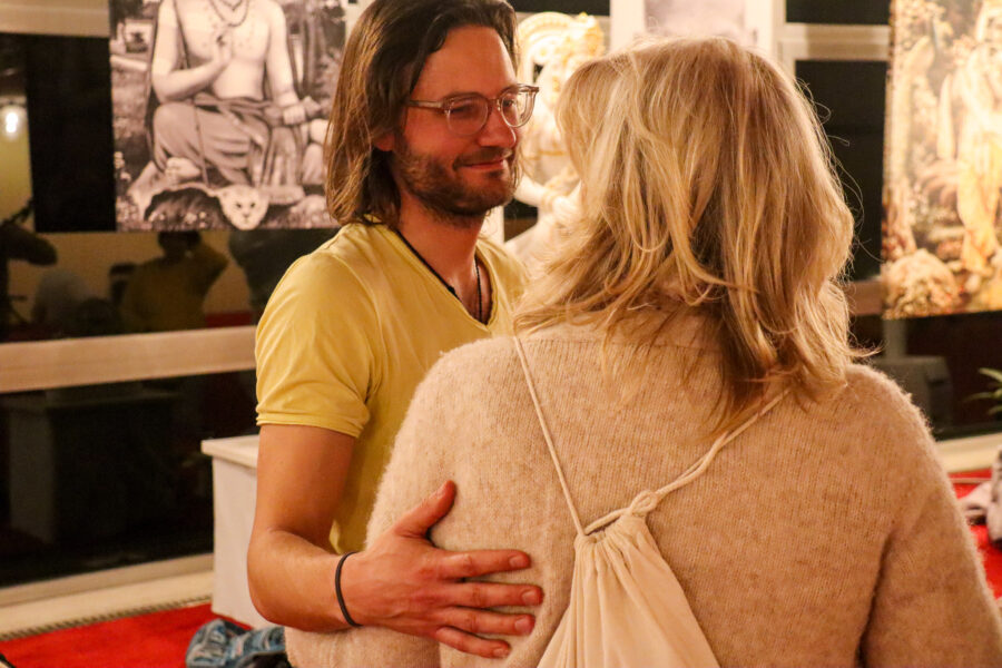 Ein Mann mit Brille und gelbem Hemd umarmt herzlich eine Frau mit blondem Haar und beigem Pullover. Sie stehen in einem Raum mit spirituellen Kunstwerken und Skulpturen im Hintergrund, die eine heitere Atmosphäre schaffen.