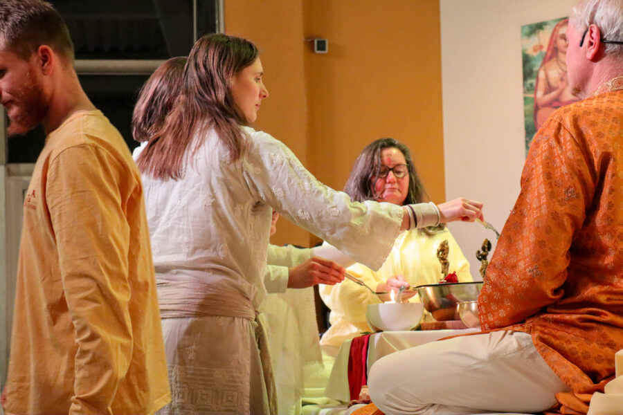 Eine Gruppe von Menschen in traditioneller Kleidung versammelt sich um einen Tisch mit spirituellen Gegenständen und kleinen Statuen. Eine Frau, die eine Brille trägt, ist aktiv an der Zeremonie beteiligt, während andere teilnehmen und zuschauen. Die Umgebung ist warm beleuchtet.