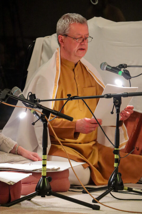 Eine Person mit Brille und traditioneller Kleidung sitzt da, hält ein Stück Papier und liest laut vor. Vor ihr sind mehrere Mikrofone aufgestellt. Ein weißes Tuch hängt über ihren Schultern und neben ihr ist eine weitere Person teilweise zu sehen.