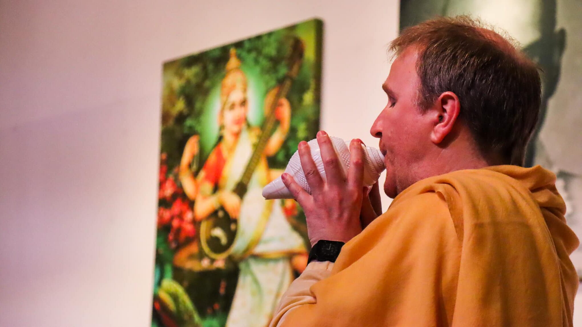 Yogalehrer bläst zur Mantraweihe in ein weißes Muschelhorn vor einem Bild der Göttin Saraswati.