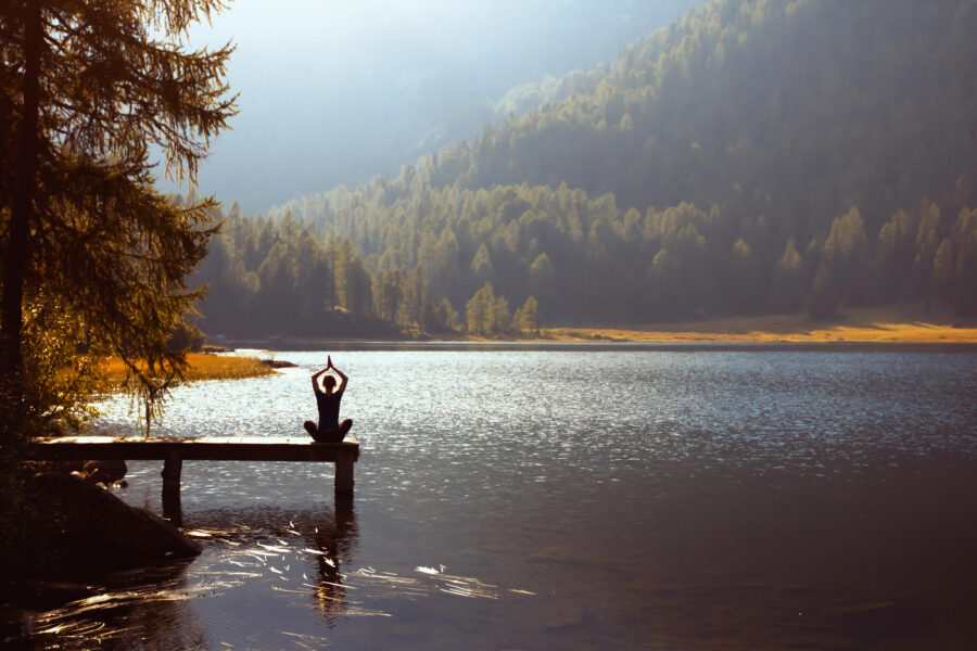 Person meditiert auf einem Steg an einem ruhigen See mit Wald und Bergen im Hintergrund